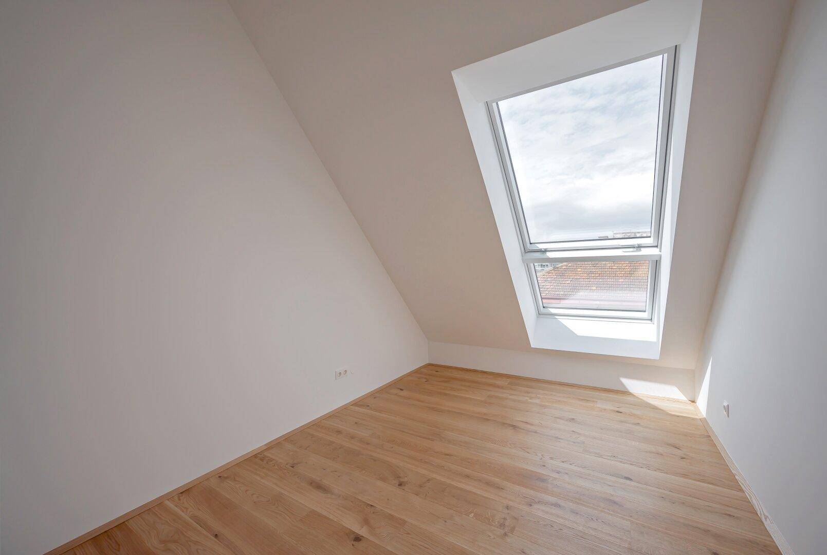 Helles Dachgeschosszimmer mit schrägem Dach und einem großen Dachfenster, das viel Licht spendet.