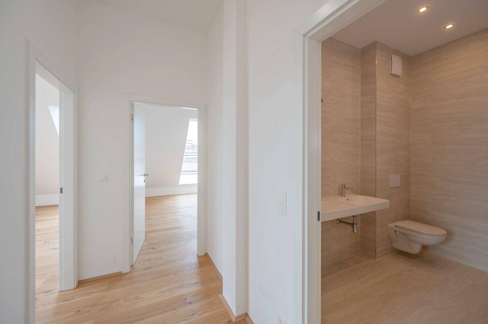 Heller Flur mit Holzboden, der zu einem modernen Badezimmer mit beigefarbenen Fliesen führt.