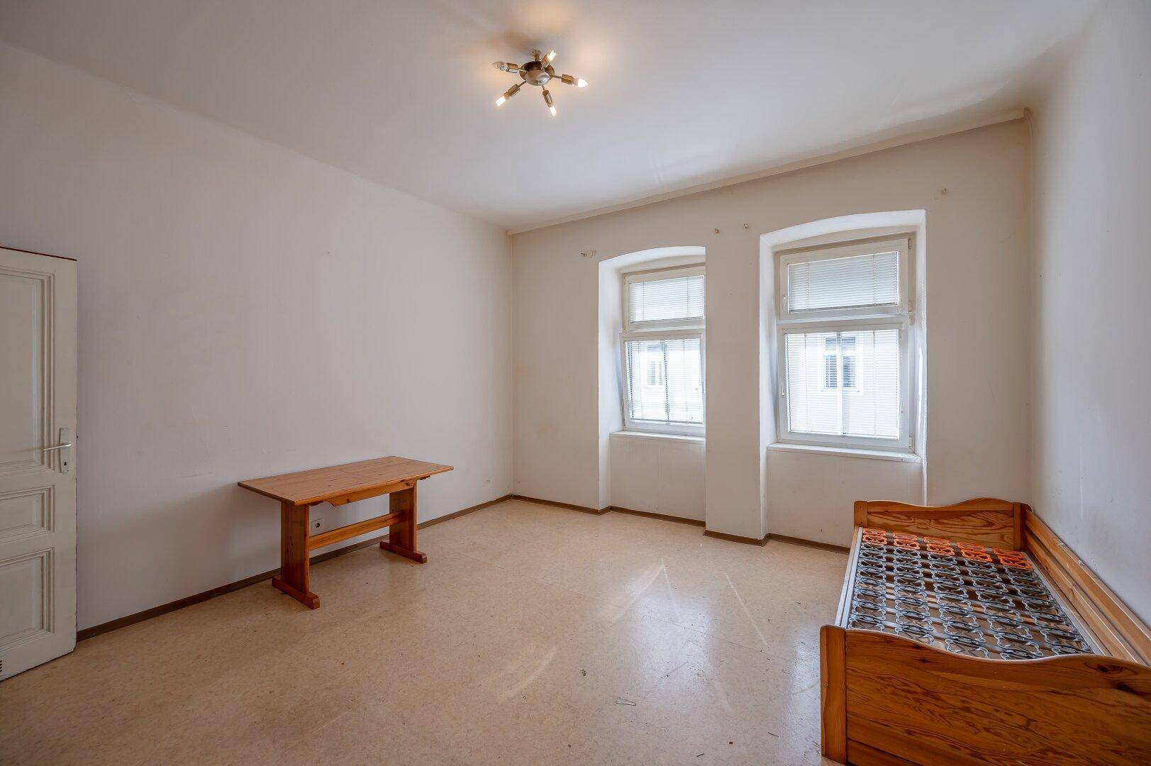 Einfaches Schlafzimmer mit Holzmöbeln, zwei Fenstern und hellem Boden, bietet Potenzial zur Renovierung.