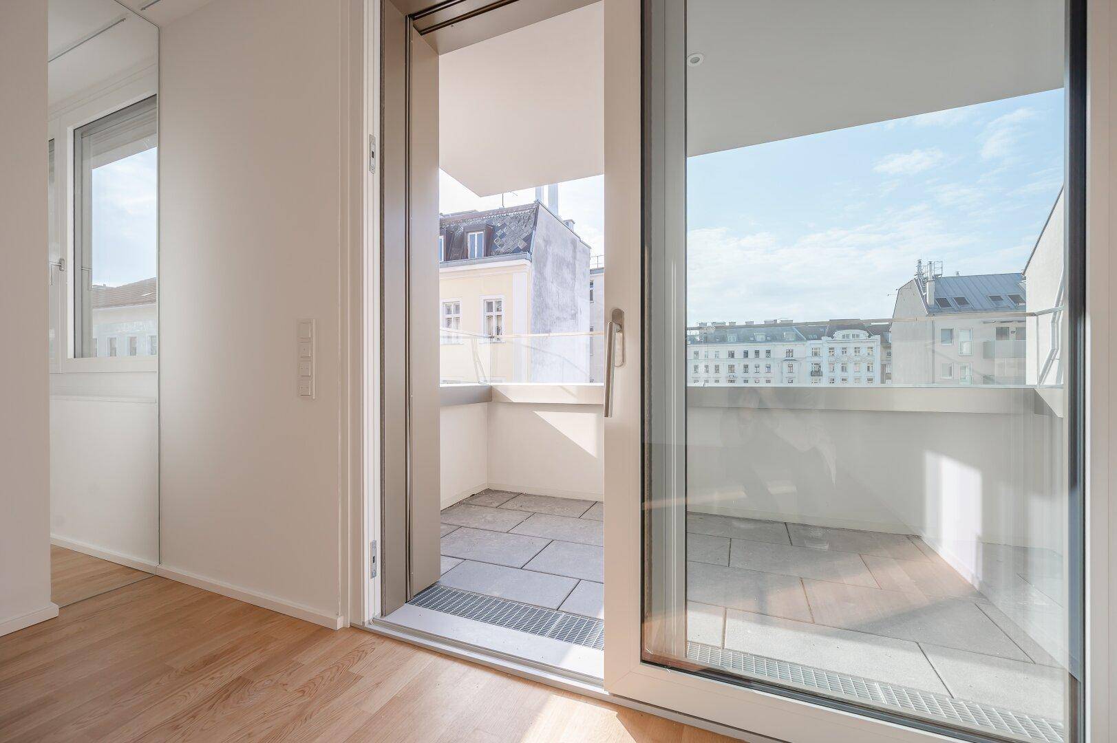 Blick aus einem hellen Innenraum durch eine große Glastür auf einen gefliesten Balkon.