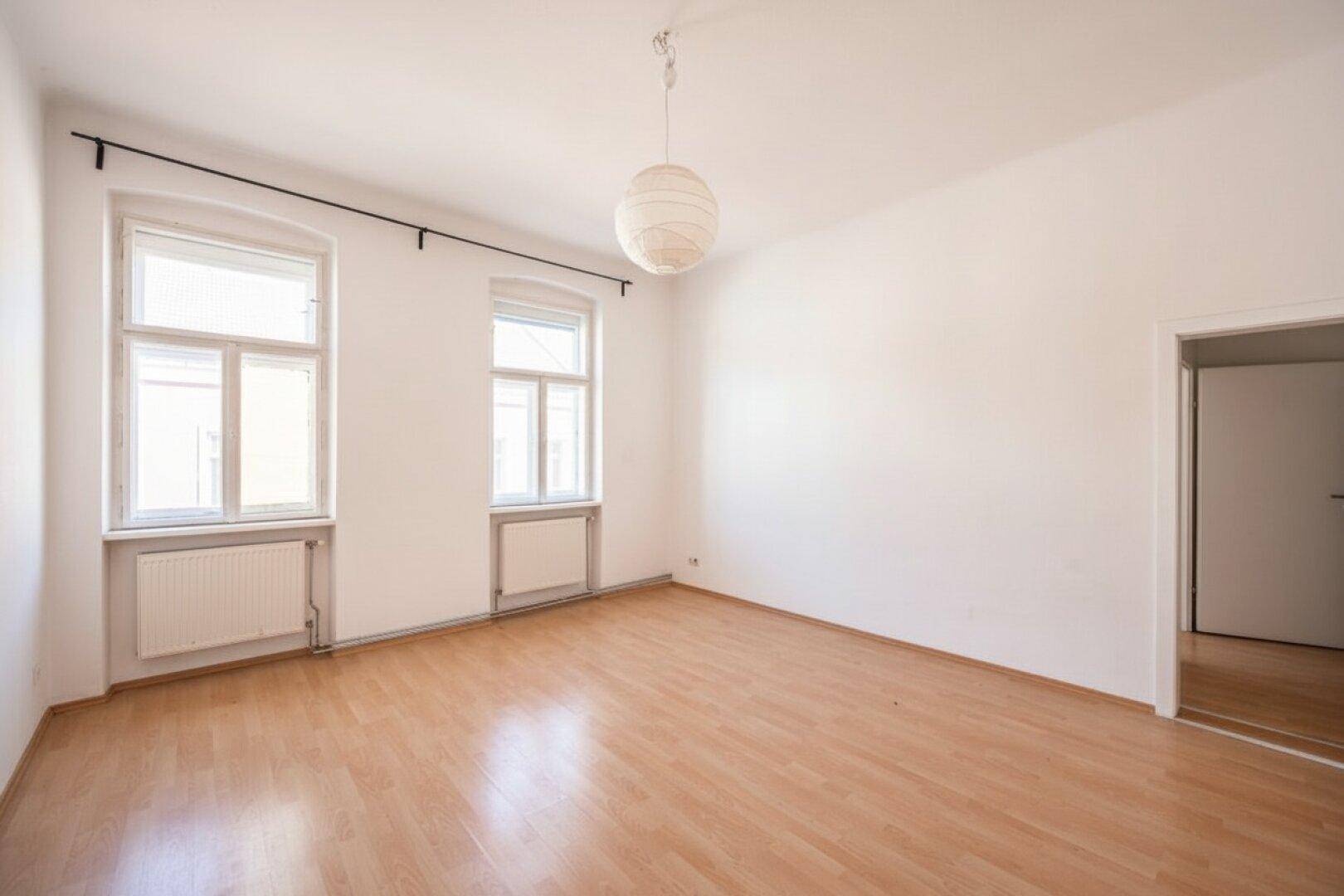 Leeres Zimmer mit hellem Holzboden, weißen Wänden und zwei Fenstern mit Heizkörpern.