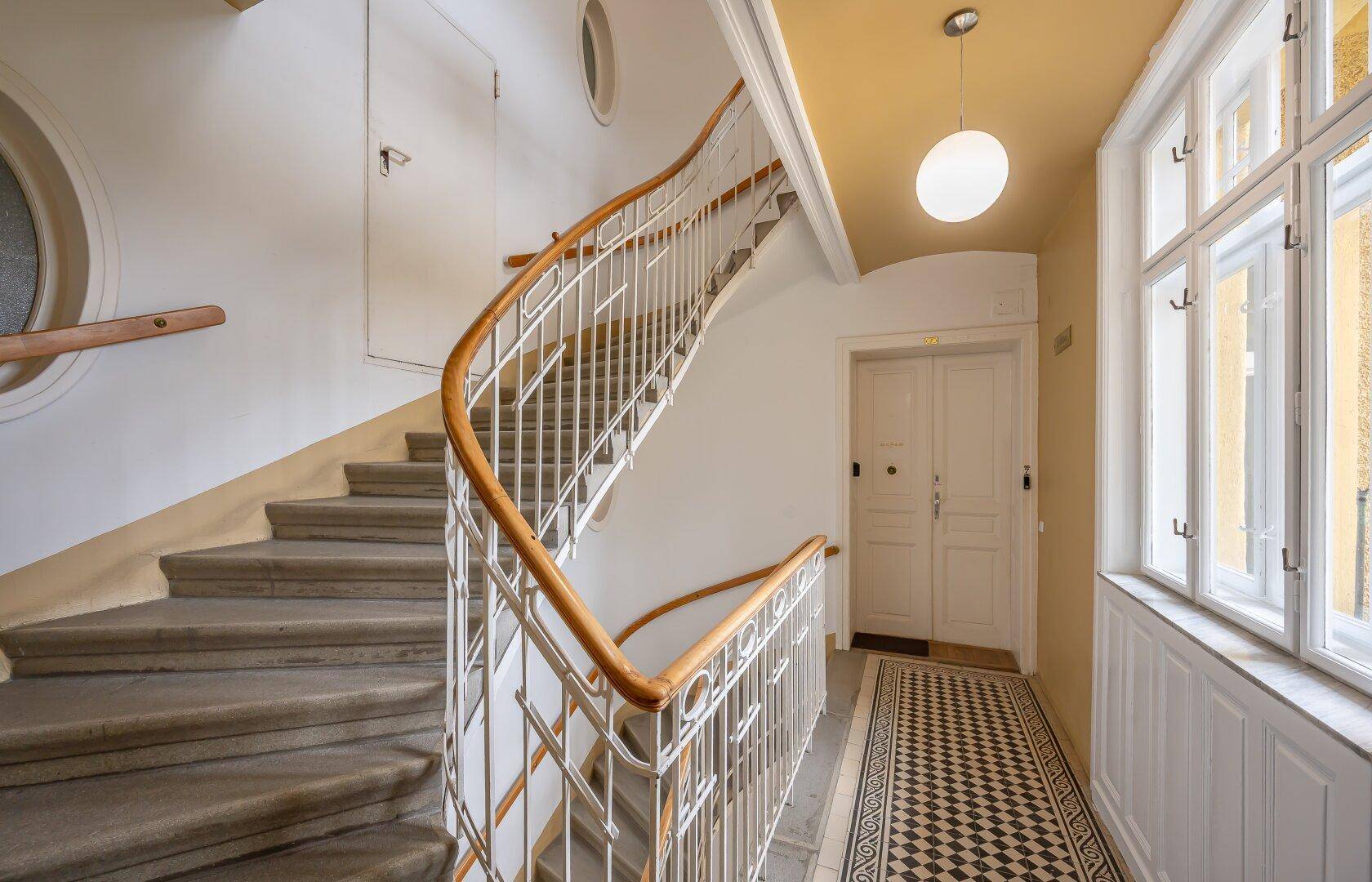 Elegantes Treppenhaus mit geschwungener Treppe, schmiedeeisernem Geländer und großen Fenstern.