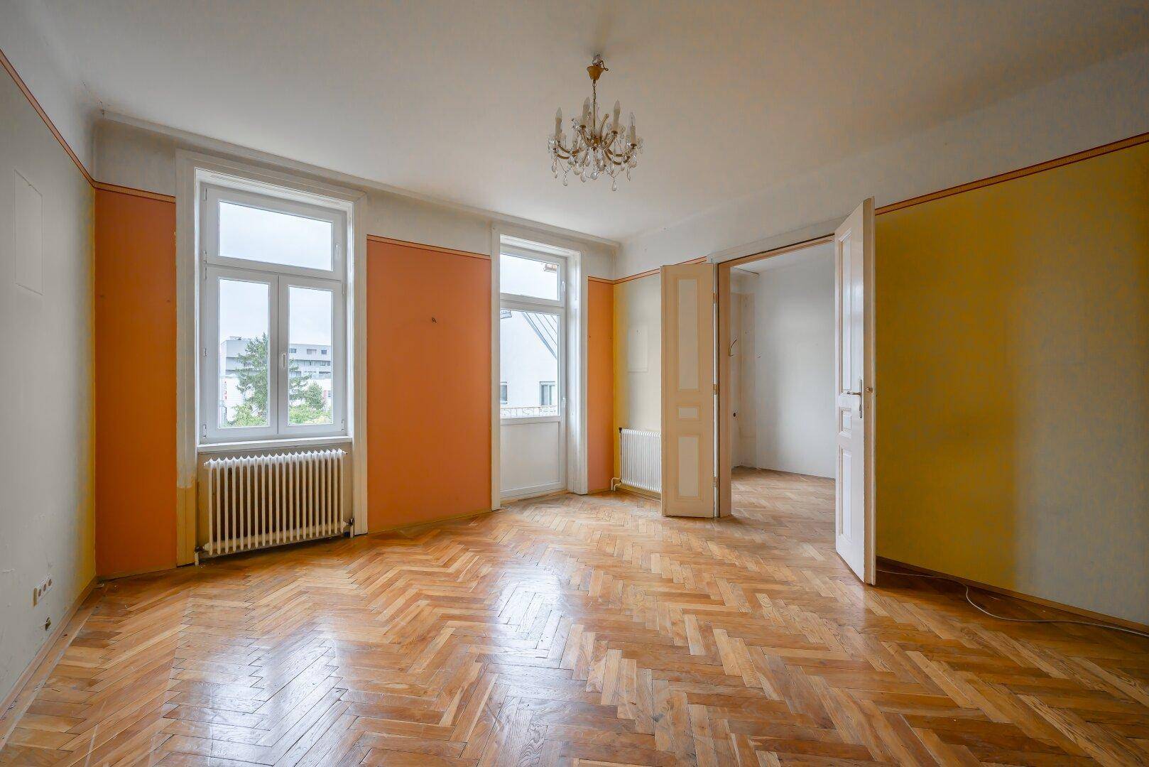 Helles Zimmer mit Fischgrätparkett, Kronleuchter und farbigen Wänden, mit Balkonzugang.