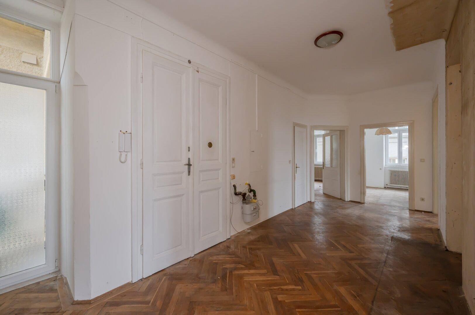 Eingangsbereich mit Fischgrätparkett, Doppeltüren und sichtbaren Installationen, bereit zur Sanierung.