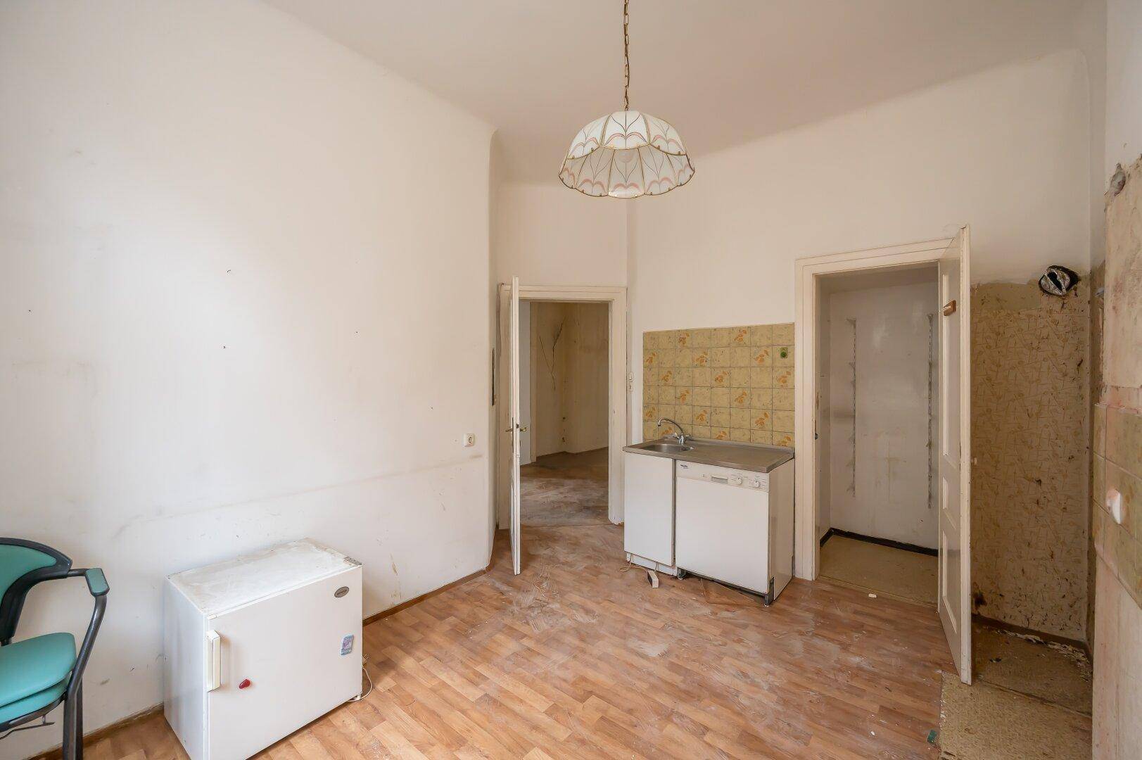Einfache Küche mit Spüle, Kühlschrank und Renovierungsbedarf, mit Zugang zu weiteren Räumen.