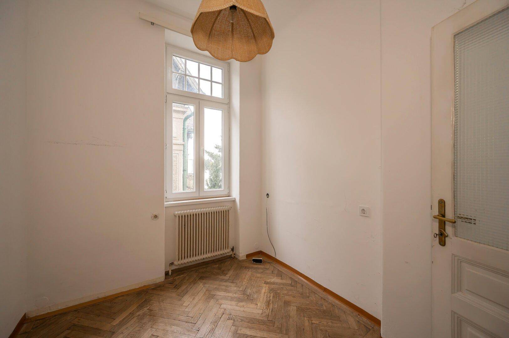 Kleines helles Zimmer mit Fischgrätparkett, großem Fenster und einfacher Deckenleuchte.