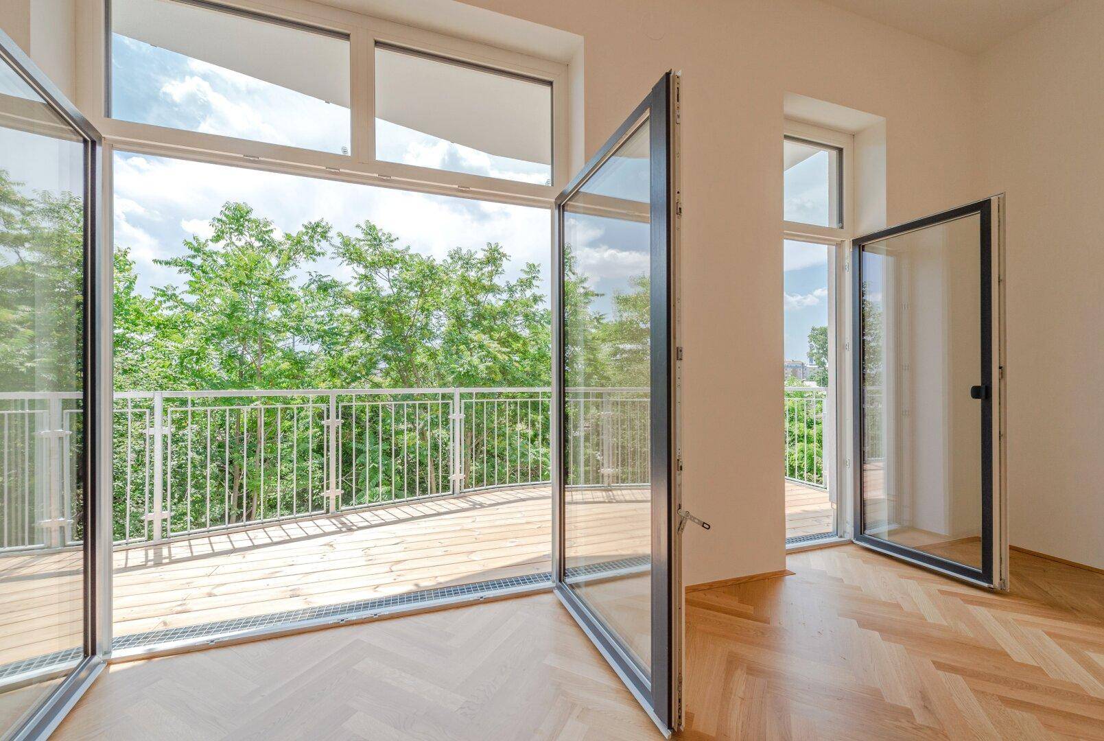 Heller Raum mit Fischgrätparkett und Zugang zu einem großen Balkon mit Blick ins Grüne.