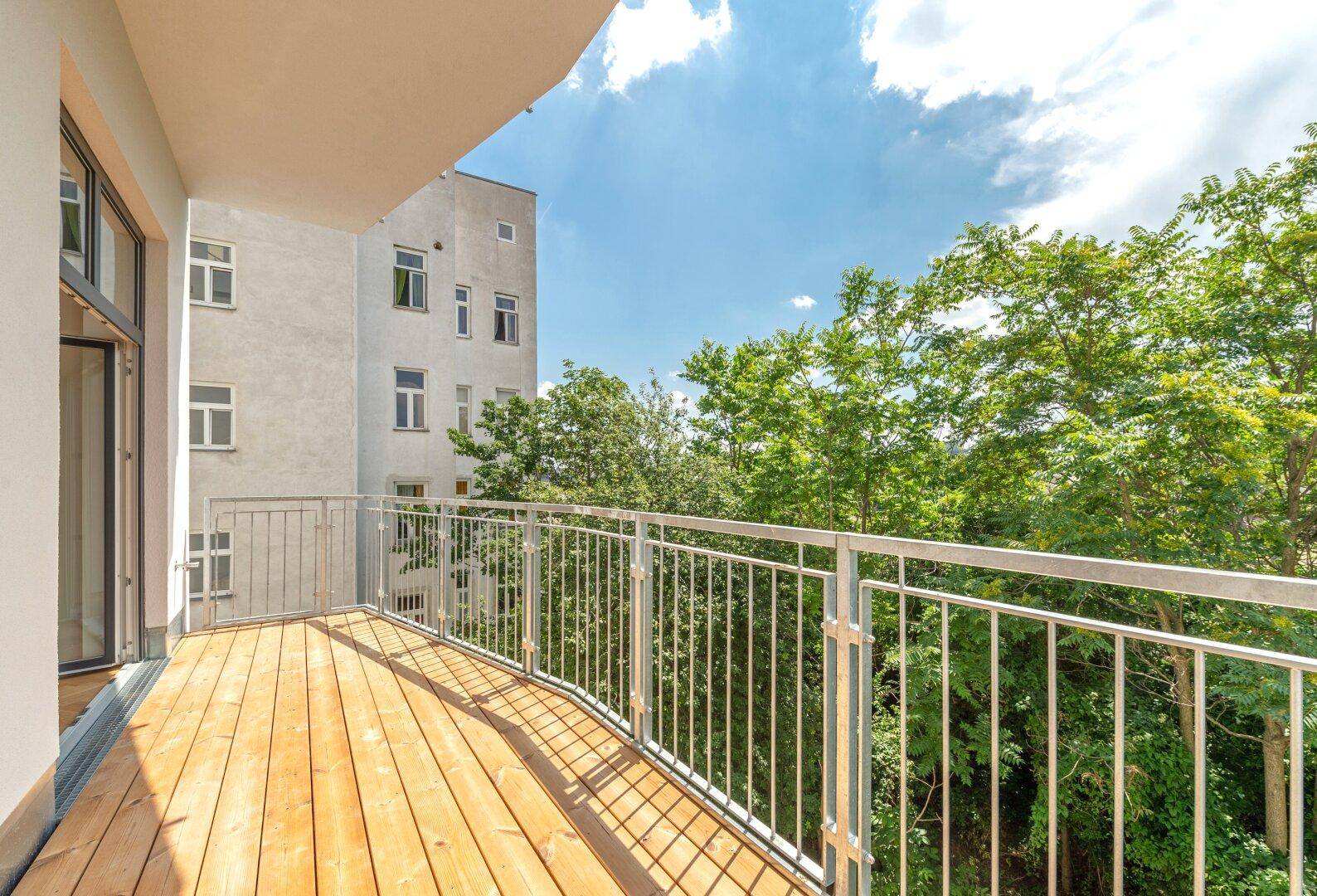 Geräumiger Holzbalkon mit Metallgeländer und Blick auf grüne Bäume und umliegende Gebäude.