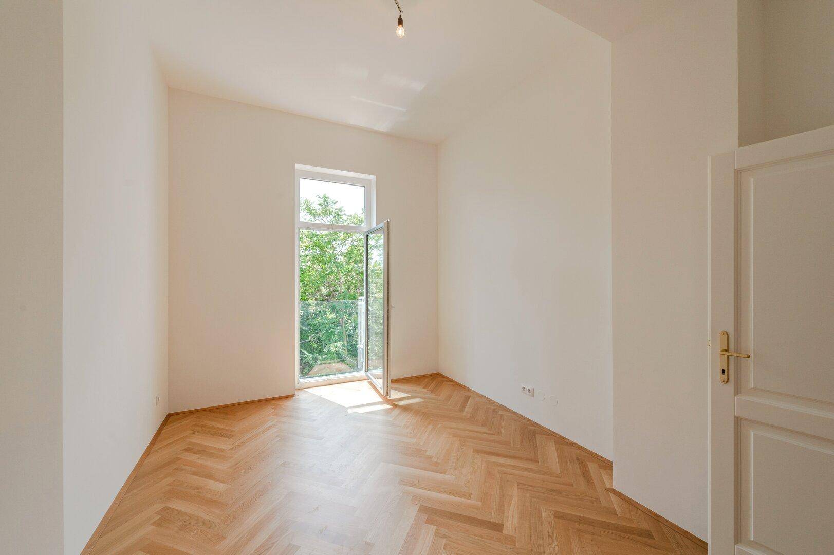 Heller, leerer Raum mit Fischgrätparkett und Fenster mit Zugang zu einem französischen Balkon.