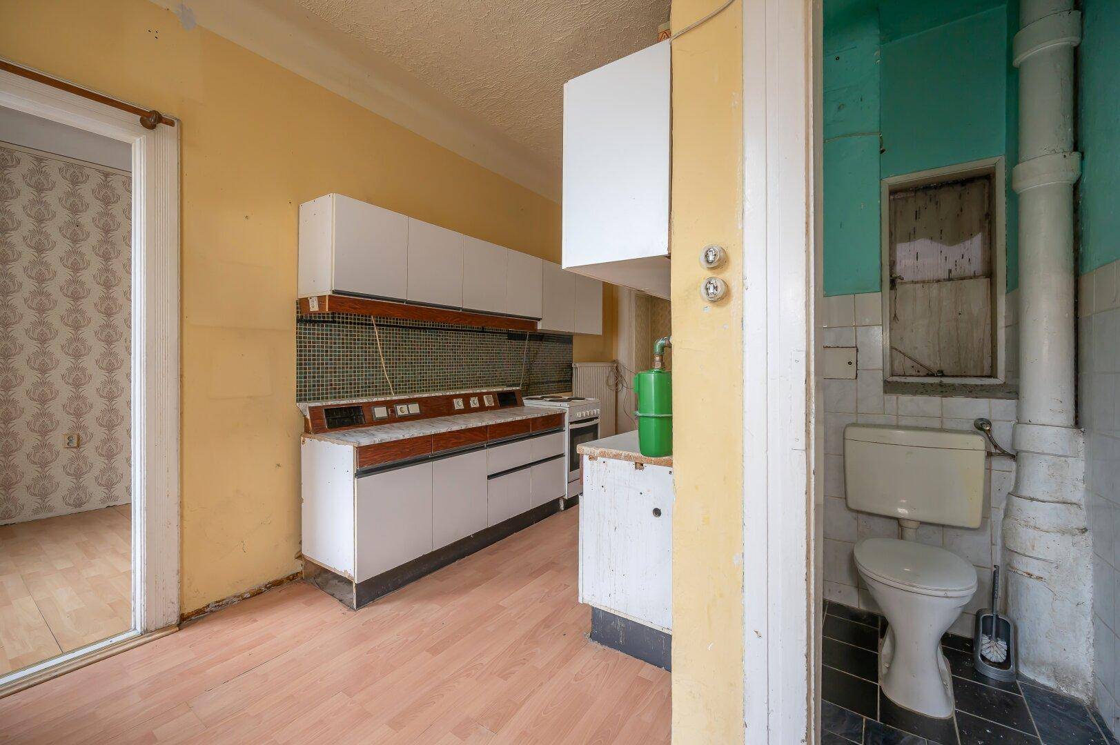 Einfache Küche mit gelben Wänden, älteren Einbauschränken und angrenzender Toilette.