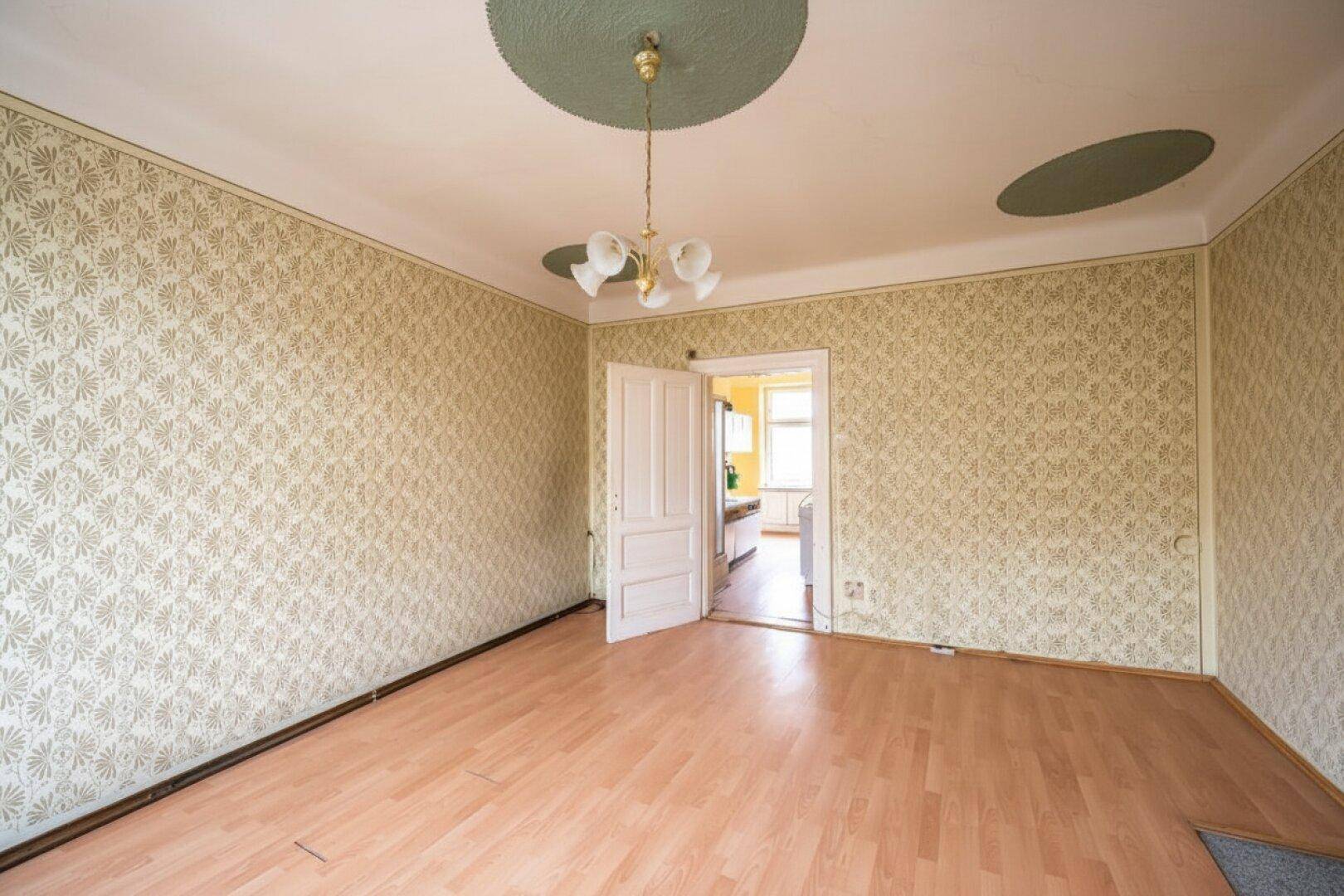 Helles Zimmer mit Laminatboden, gemusterter Tapete und Zugang zu einem weiteren Raum.
