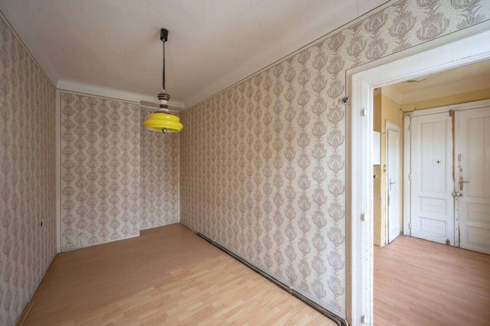 Leeres Zimmer mit Laminatboden, gemusterter Tapete und gelber Deckenleuchte.