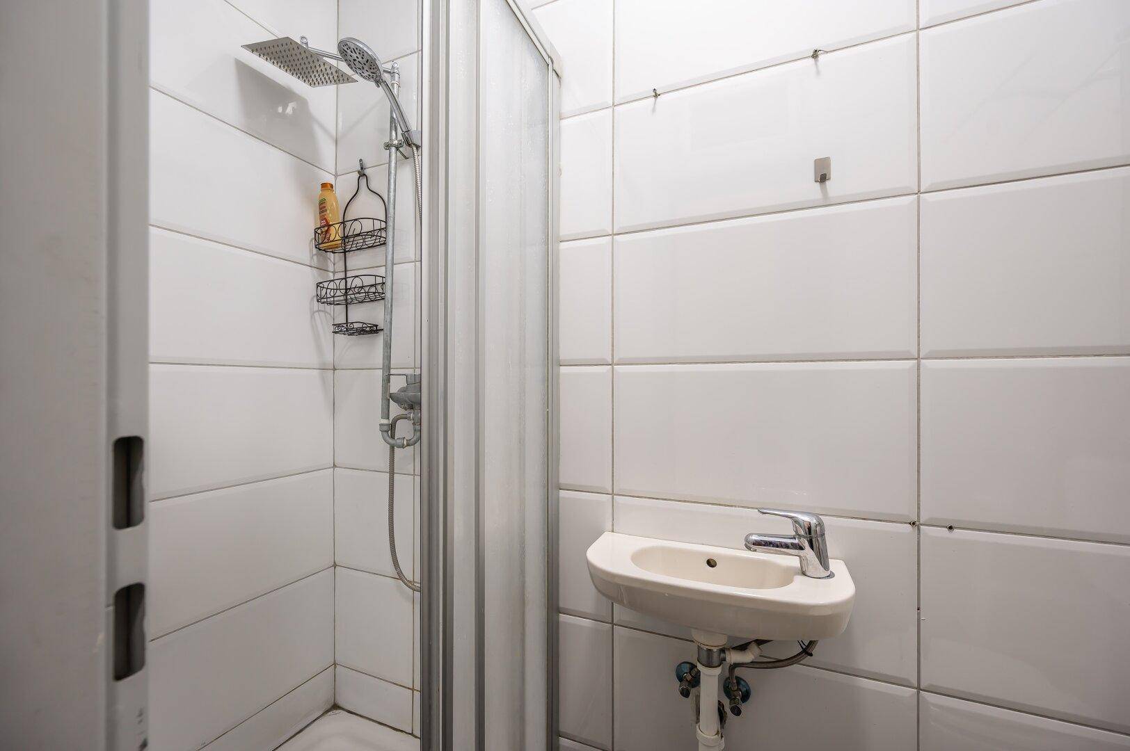 Kompaktes Badezimmer mit Duschkabine, kleinem Waschbecken und weißen Wandfliesen.