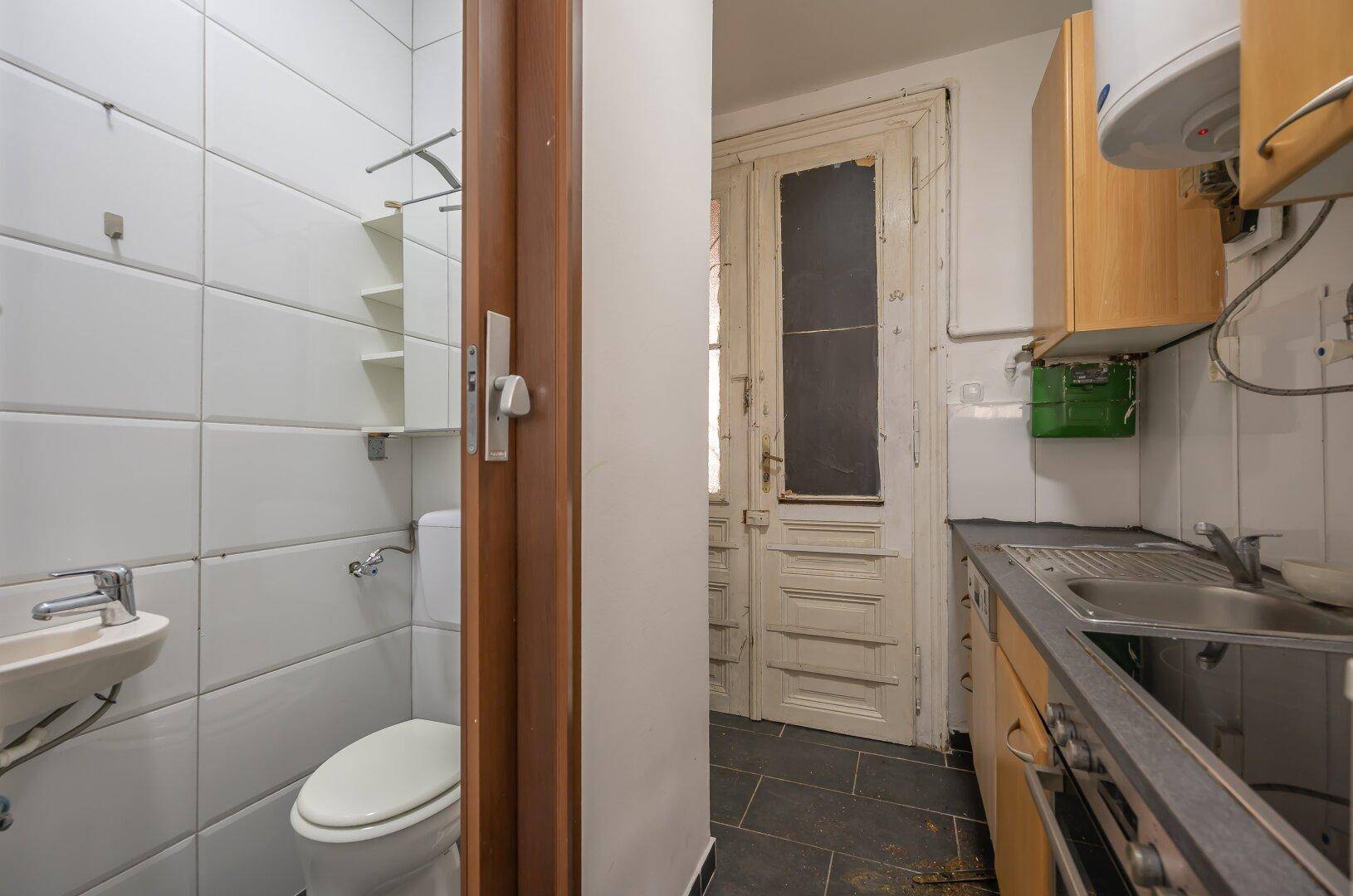 Kleines WC mit Waschbecken und Zugang zu einer Küche, gekennzeichnet durch eine abgenutzte Holztür.