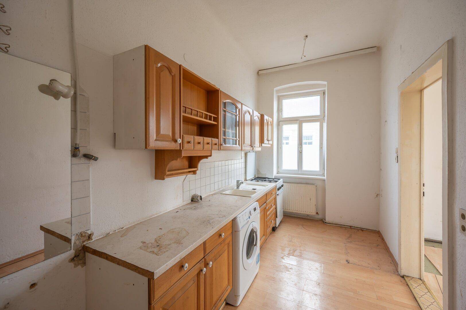 Renovierungsbedürftige Küche mit älteren Holzschränken, Spüle, Herd und Waschmaschine unter dem Fenster.
