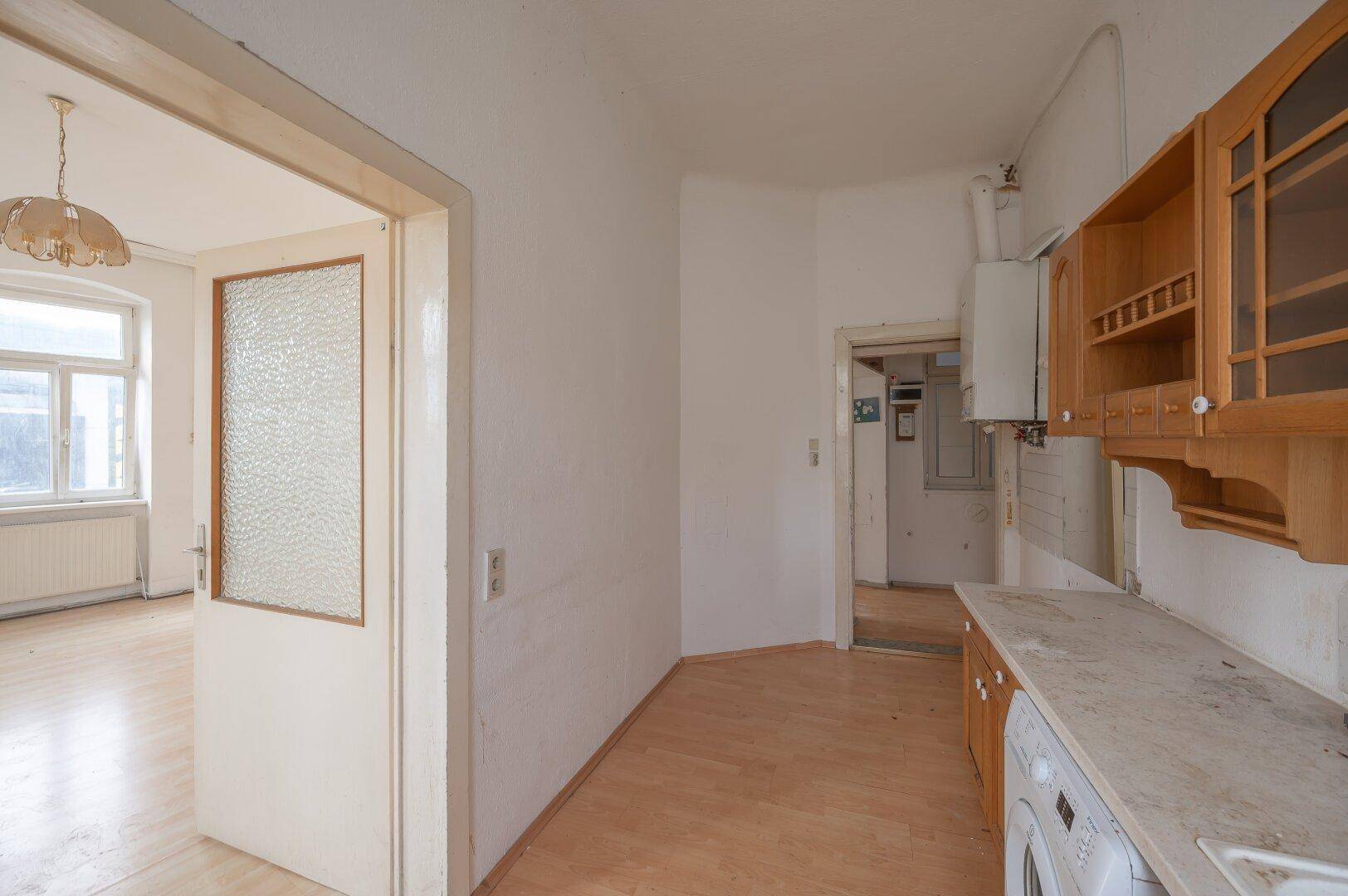 Küche mit älteren Holzschränken und Durchgang zu einem weiteren Zimmer, renovierungsbedürftig.