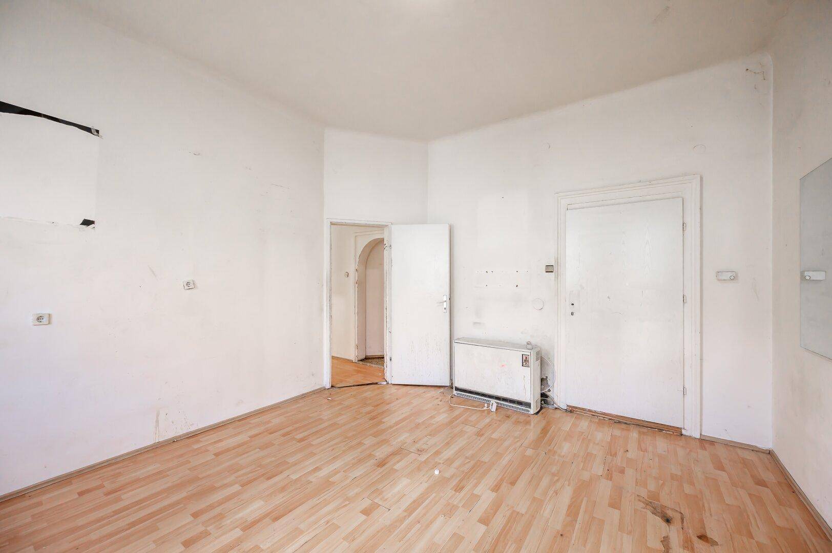 Leeres Zimmer mit Laminatboden, Heizkörper und mehreren Türen, das eine Renovierung benötigt.