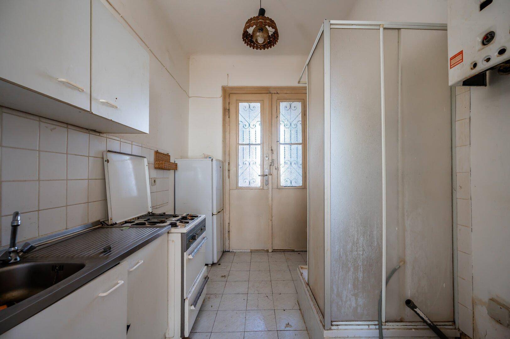 Veraltete Küche mit Fliesenboden, Spüle, Gasherd, Kühlschrank und einer Duschkabine.