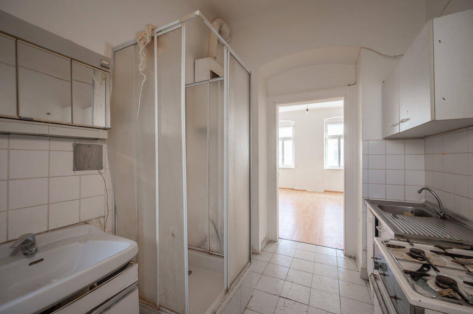 Abgenutzte Küche mit integrierter Duschkabine und Blick in ein angrenzendes Zimmer.