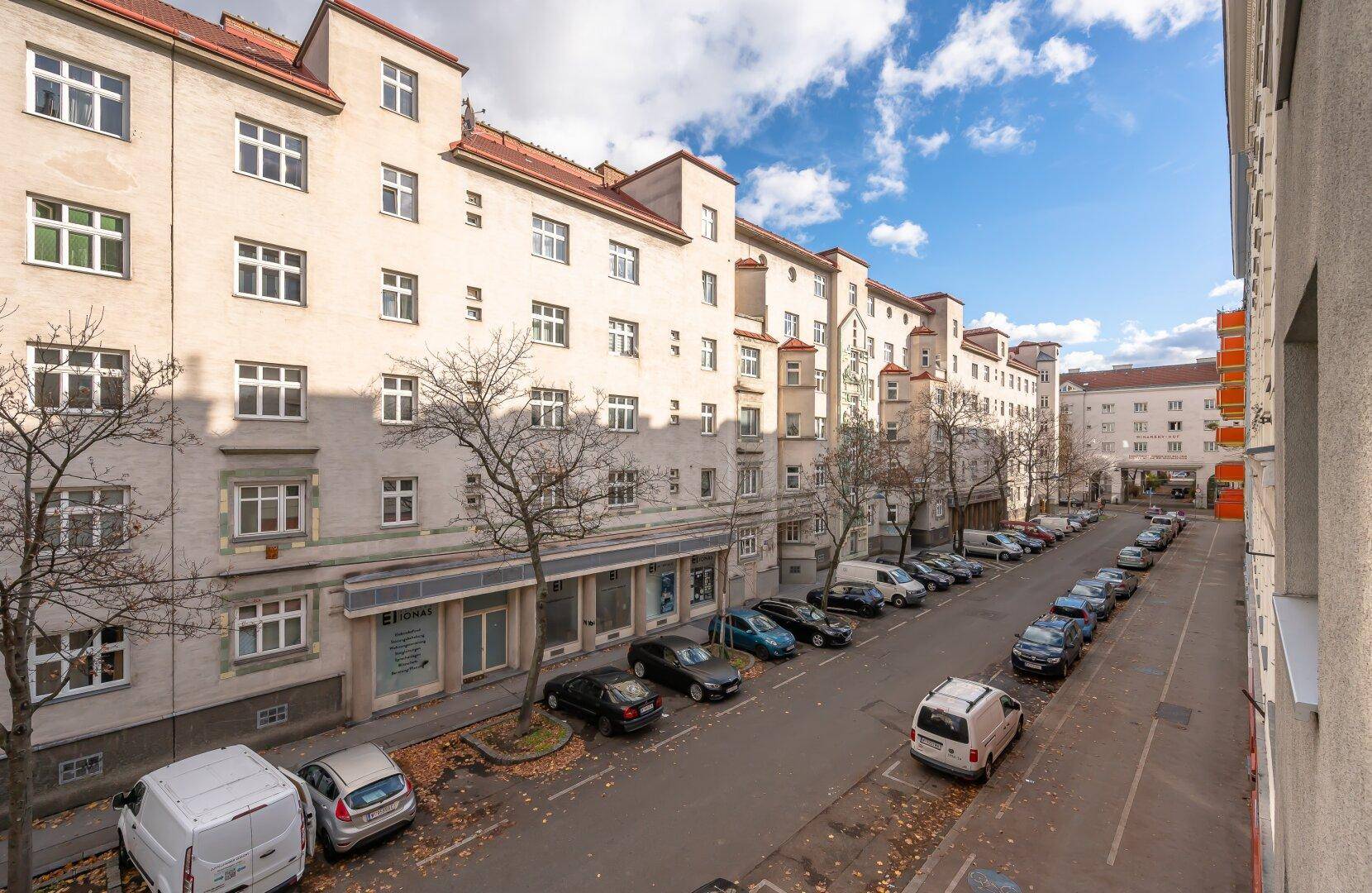 Lange Wohnstraße mit parkenden Fahrzeugen und klassischen Gebäudefassaden unter blauem Himmel.