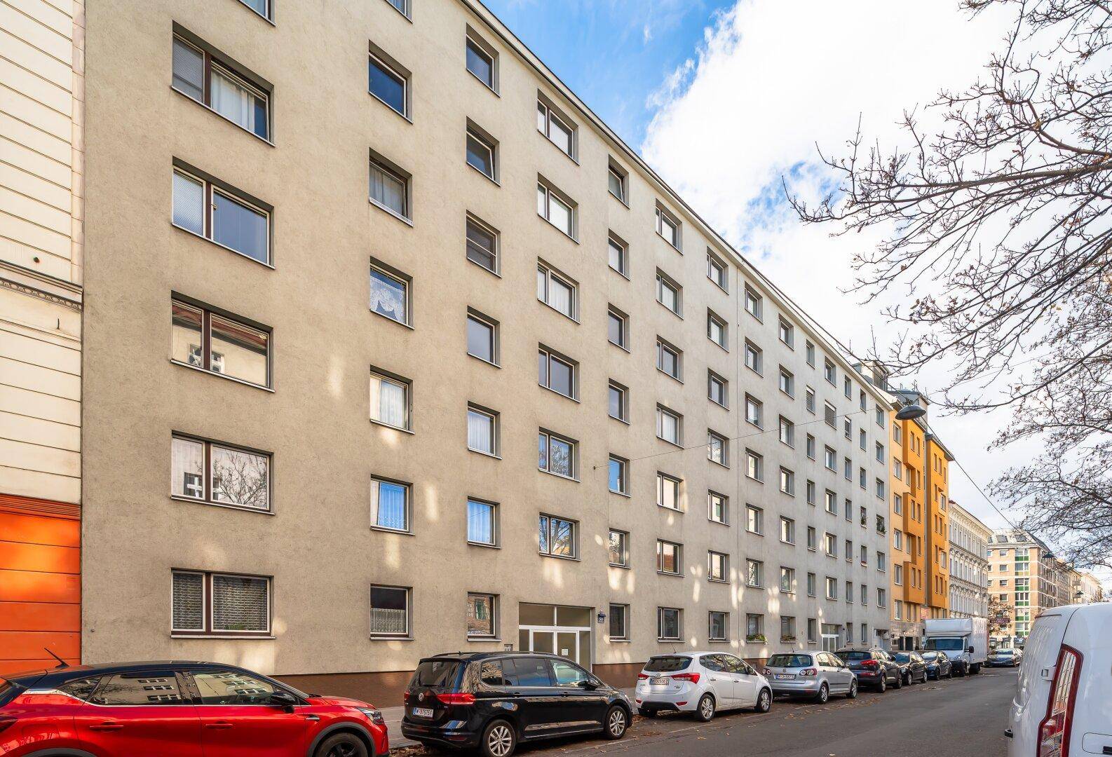 Lange Fassade eines Wohngebäudes mit vielen Fenstern und parkenden Autos auf der Straße.