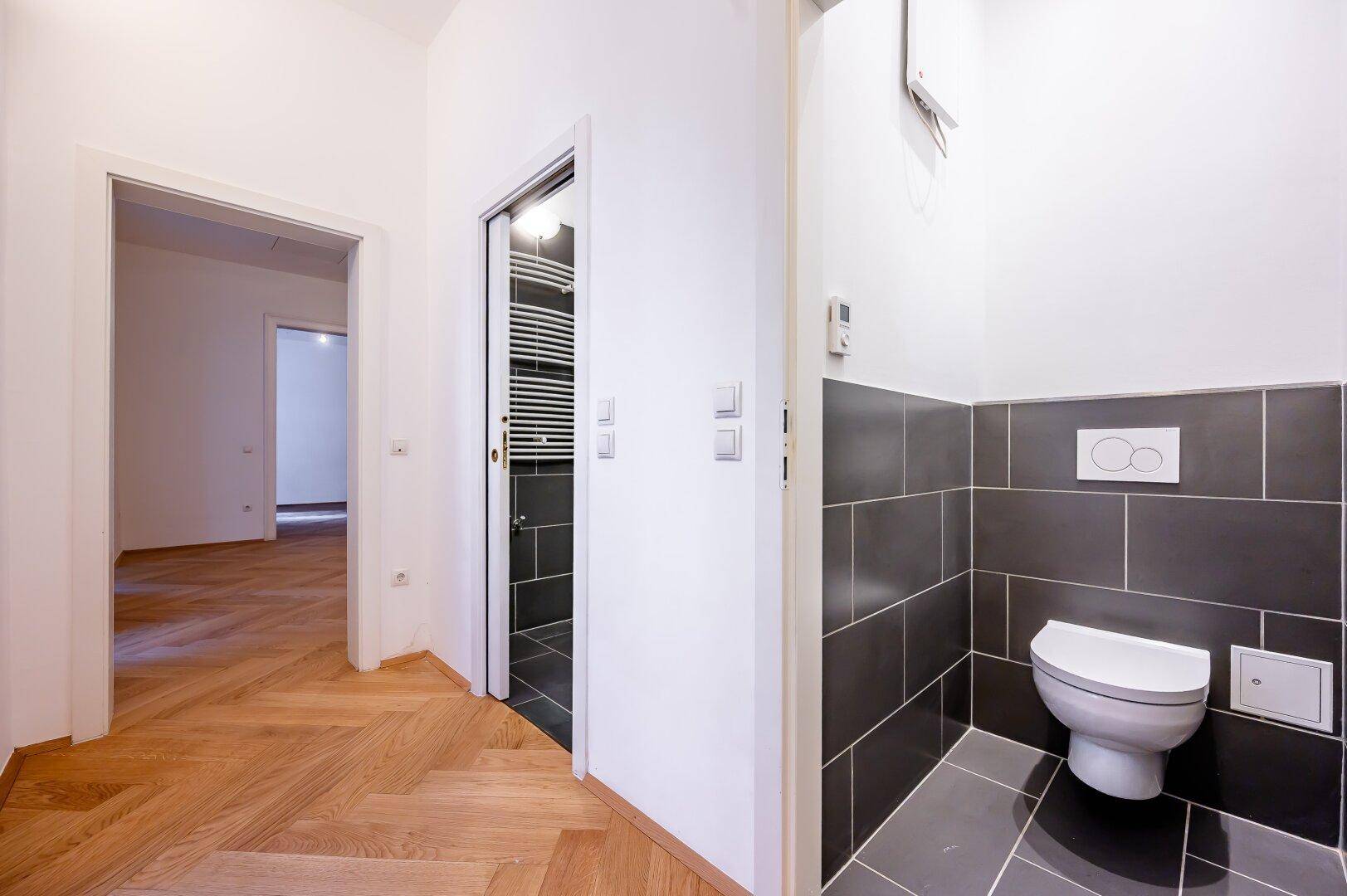 Flur mit Fischgrätparkett und Zugang zu einem modernen Gäste-WC mit dunklen Fliesen.