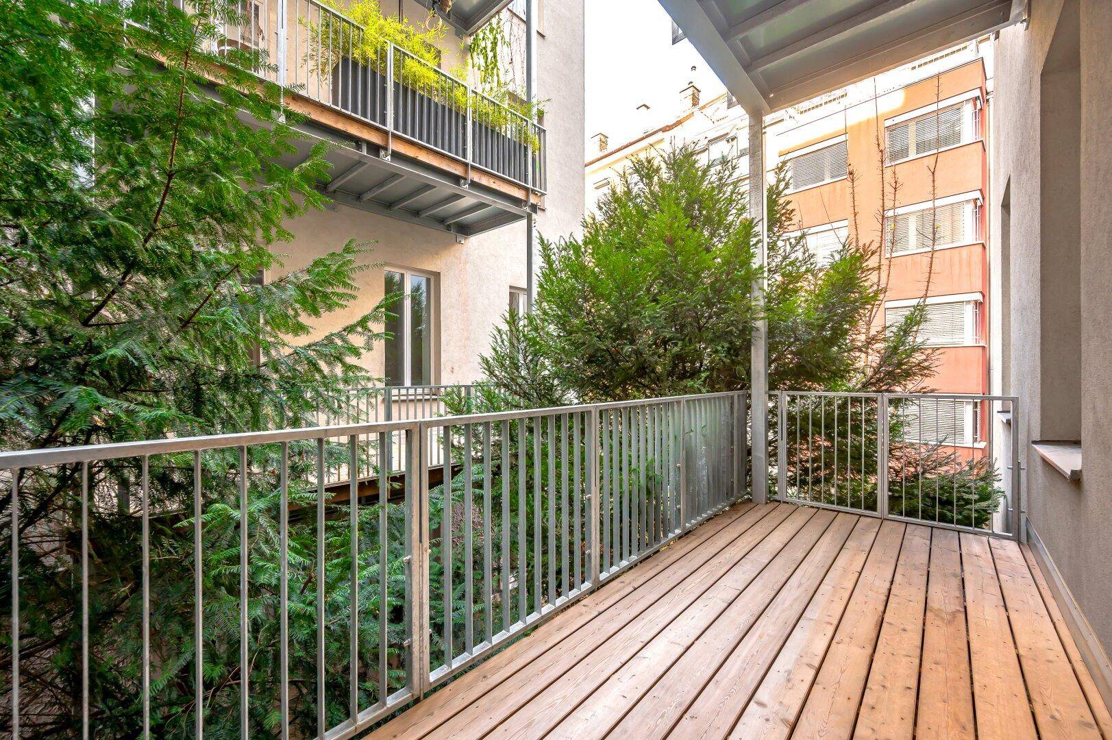 Geräumiger Balkon mit Holzfußboden und Metallgeländer, umgeben von grünen Bäumen.