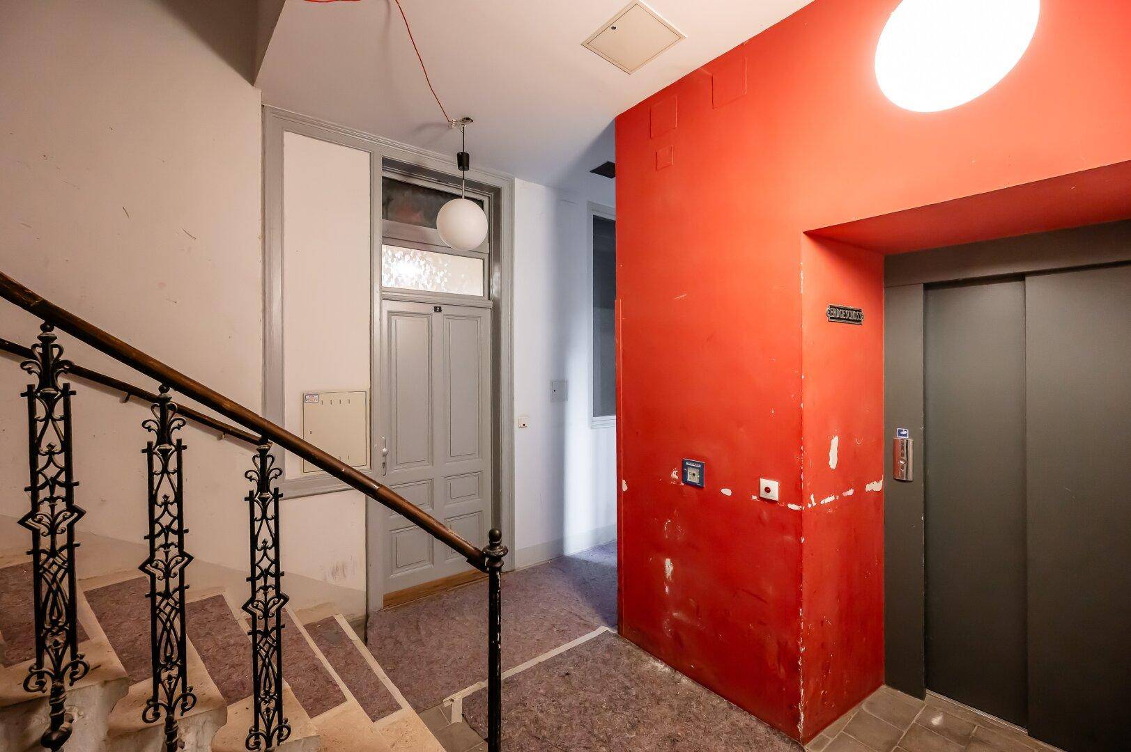 Eingangsbereich mit Treppenhaus, historischem Geländer und modernem Aufzug in roter Wand.