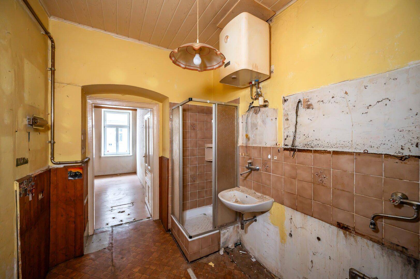 Badezimmer in sanierungsbedürftigem Zustand mit Duschkabine, Waschbecken und Warmwasserboiler.