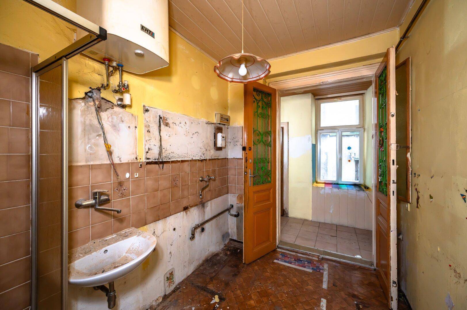 Sanierungsbedürftiges Badezimmer mit Duschkabine, Waschbecken und sichtbaren Installationen.