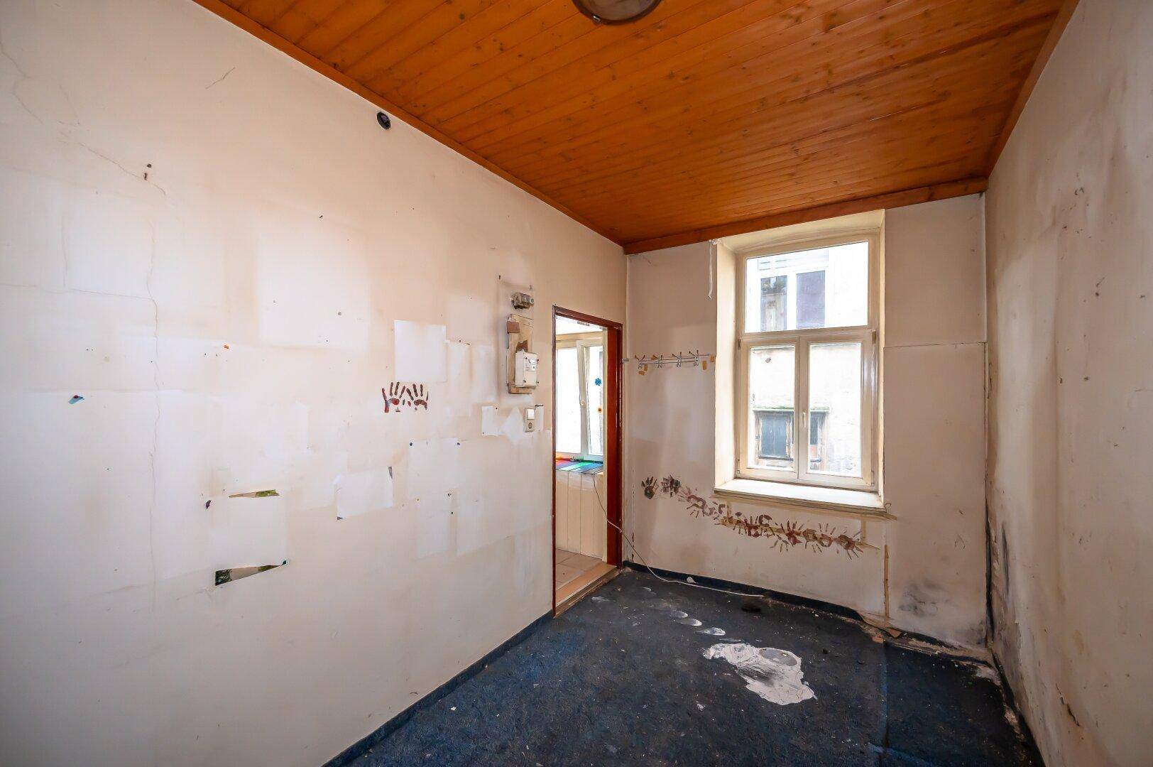 Kleiner Raum mit Teppichboden, Holzdecke und einem Fenster, das Tageslicht hereinlässt.