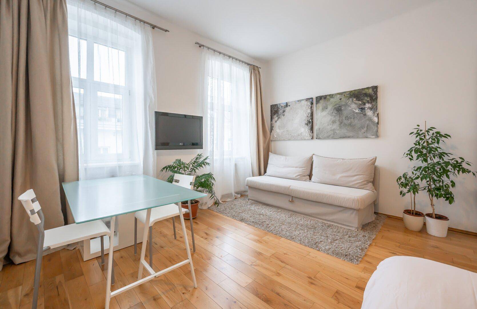 Heller Wohnbereich mit Holzboden, beigem Sofa, Esstisch und Deckenventilator, gut beleuchtet durch zwei Fenster.