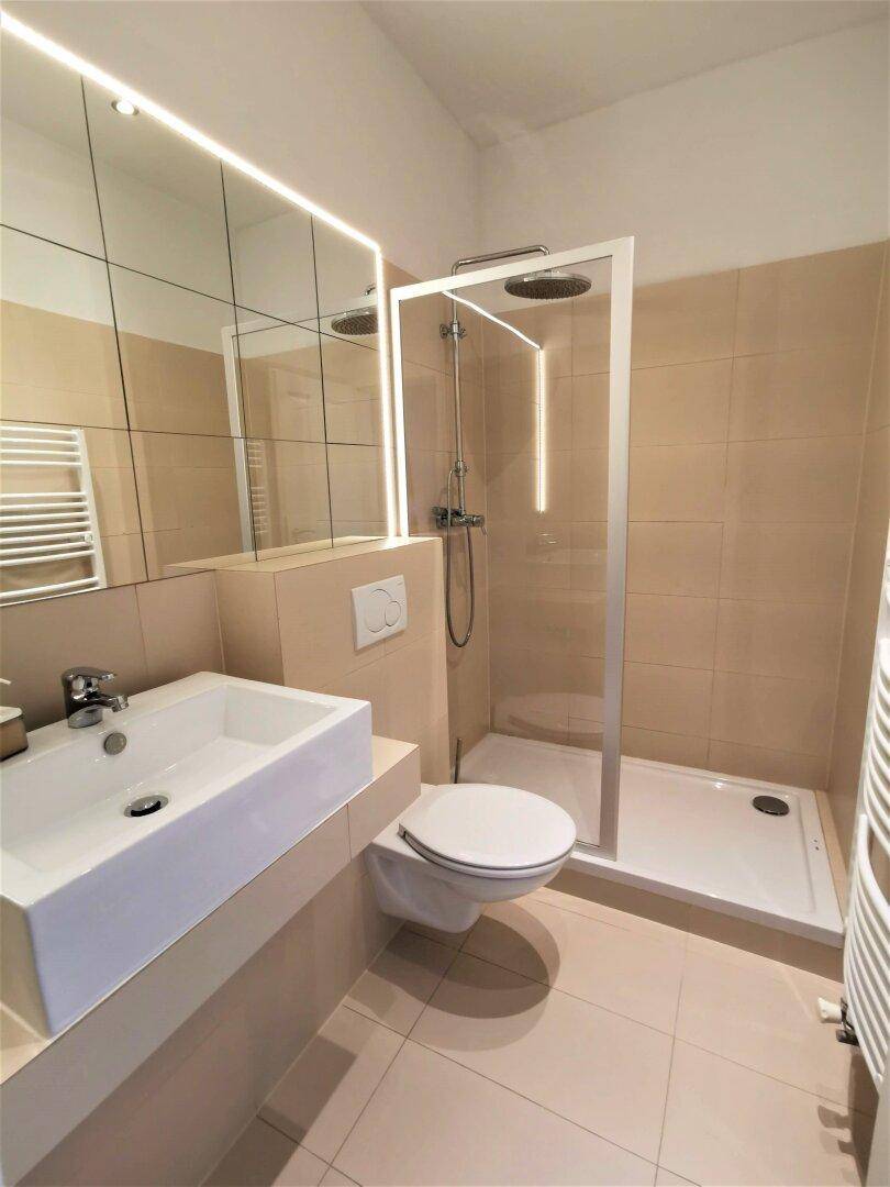Modernes Badezimmer mit beigen Fliesen, ebenerdiger Dusche, wandhängendem WC und beleuchtetem Spiegel.