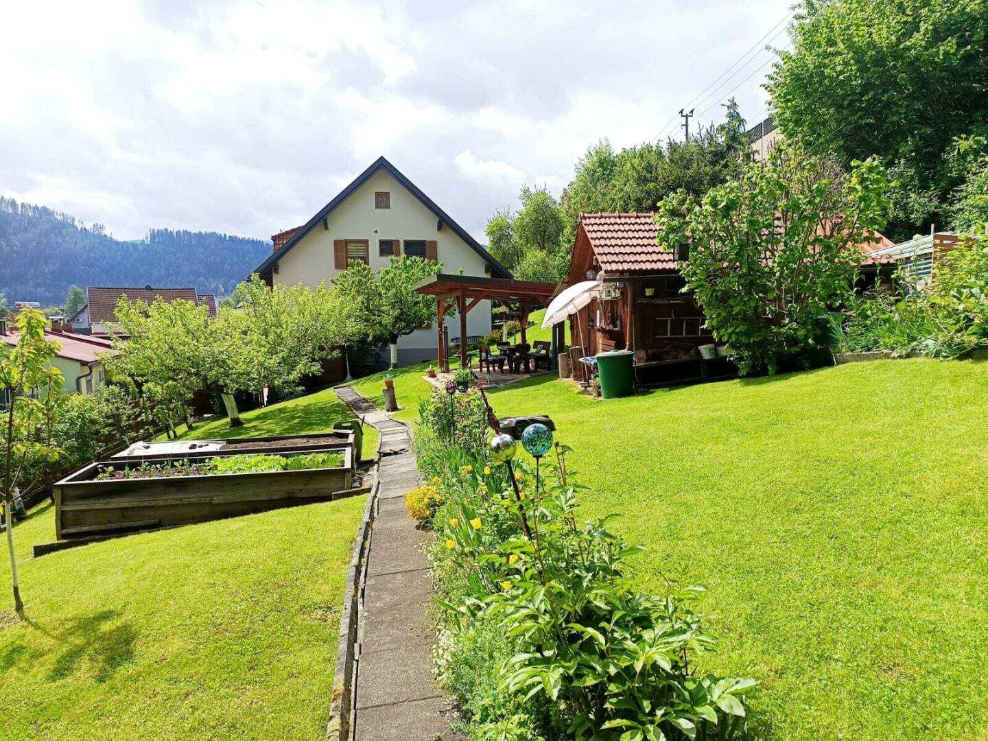 Großer, gepflegter Garten mit Rasenflächen, Hochbeeten und überdachtem Sitzbereich vor dem Haus.