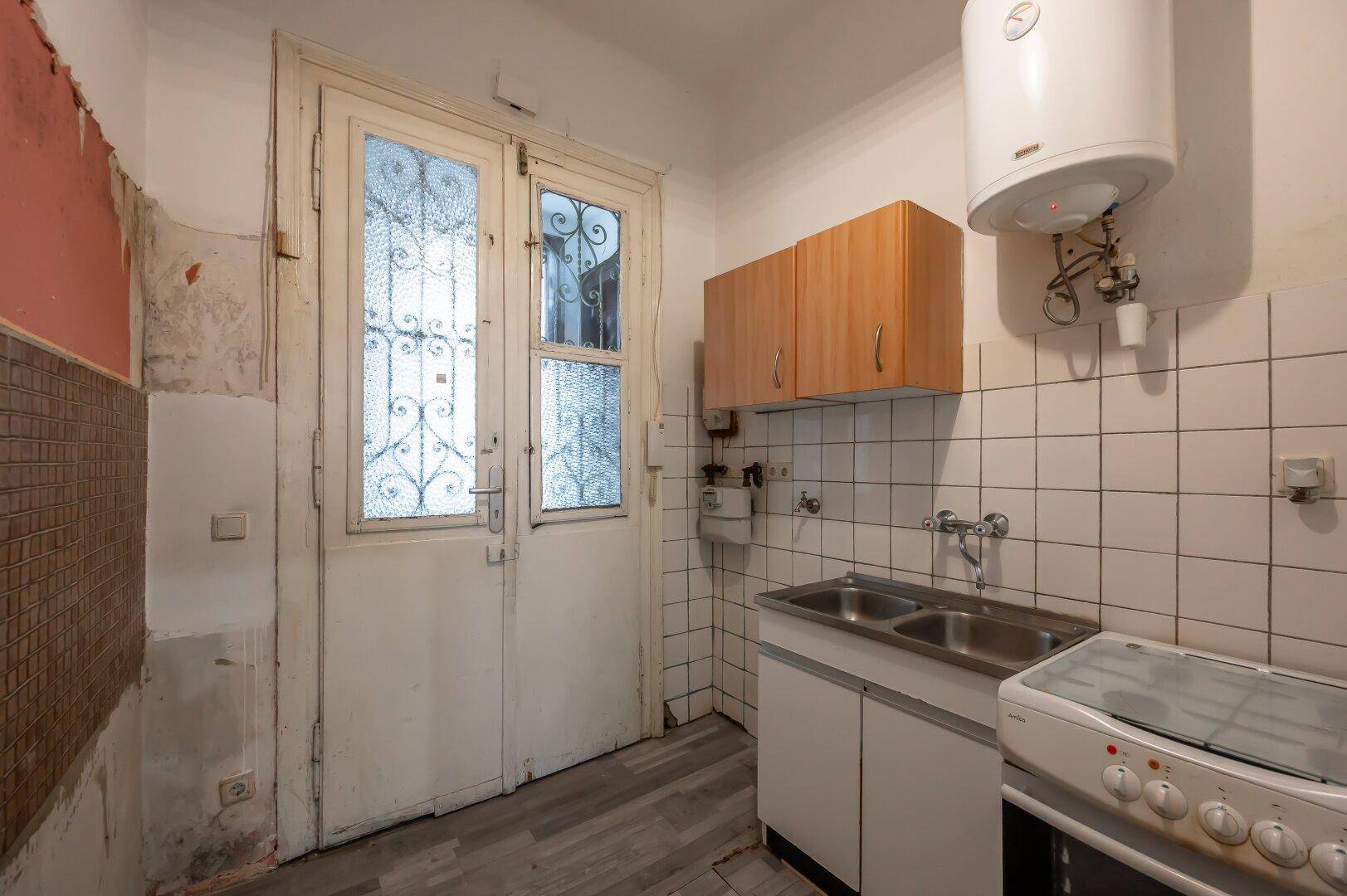 Kleine Küche mit Spüle, Herd, Hängeschränken und Boiler, renovierungsbedürftig.