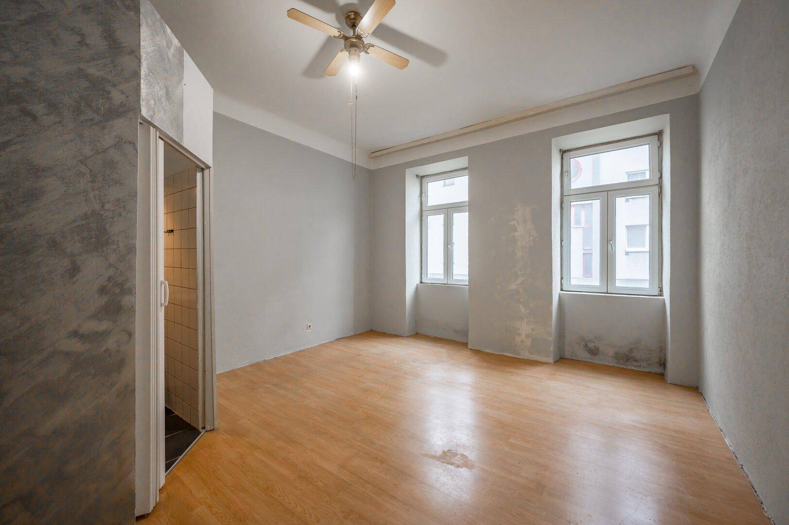 Heller Innenraum mit zwei Fenstern, Holzfußboden und Deckenventilator, bereit für die Renovierung.