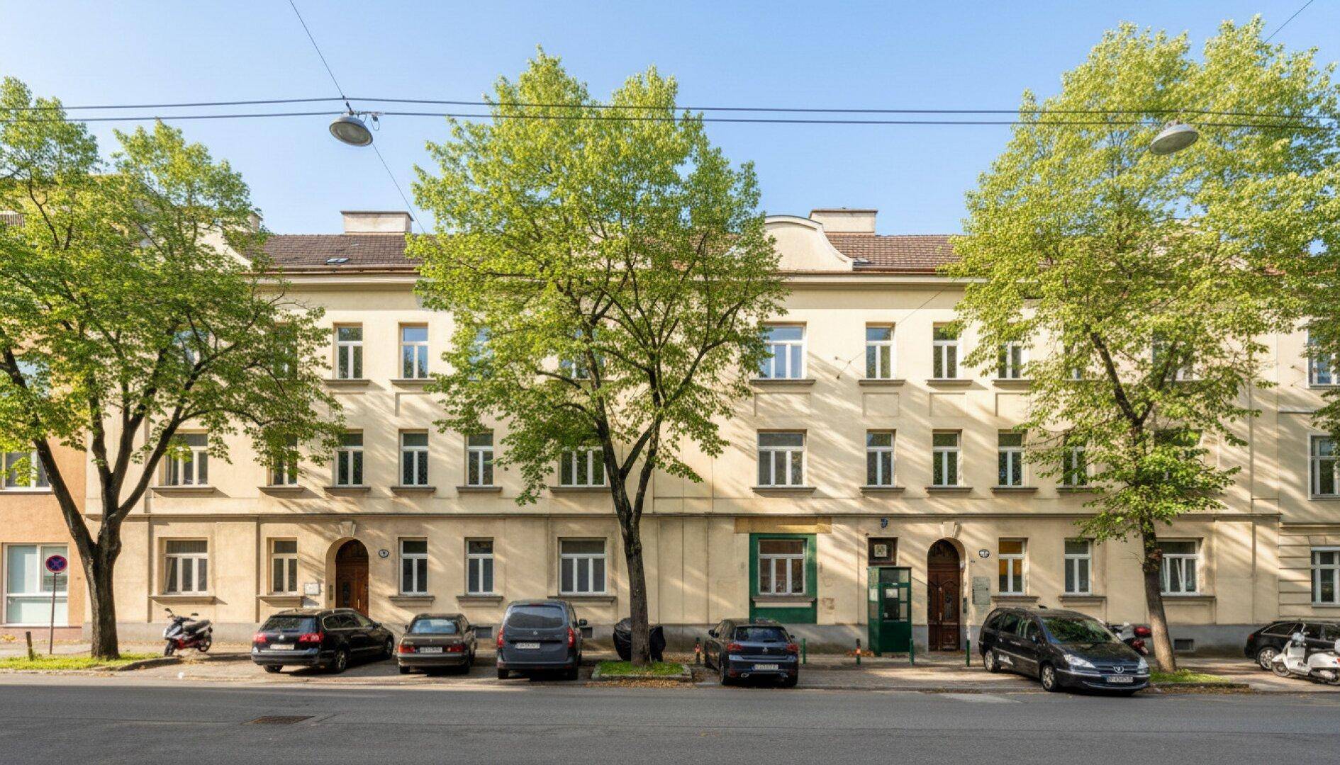 Klassische Fassade eines Mehrfamilienhauses mit vielen Fenstern und grünen Bäumen davor.