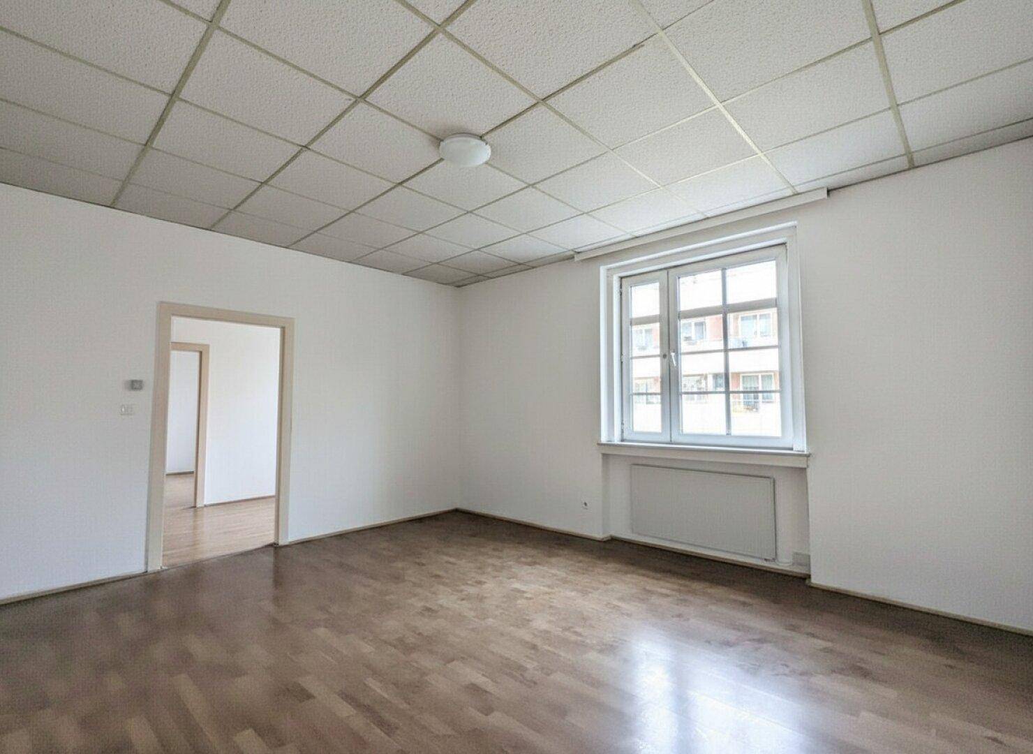 Geräumiges Zimmer mit Parkettboden, Fenster und einer Decke mit Akustikplatten.