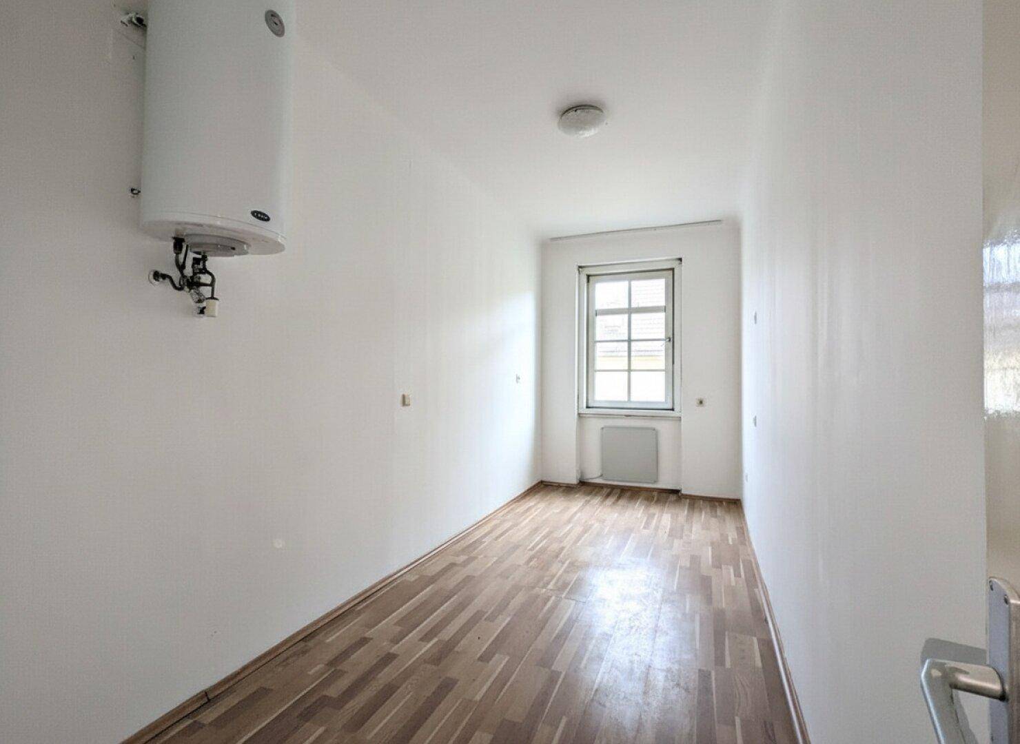 Schmaler Raum mit Holzfußboden, Fenster und einem an der Wand montierten Warmwasserbereiter.