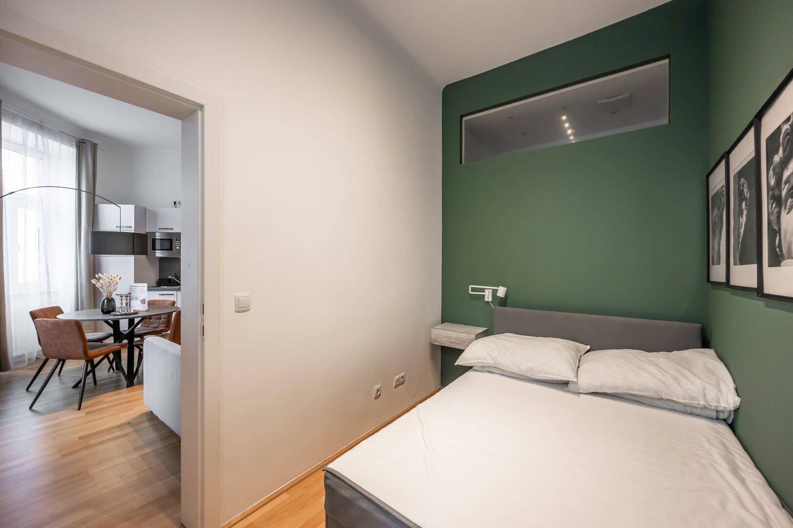 Schlafzimmer mit grüner Akzentwand, Bett und Verbindung zum Wohnbereich.