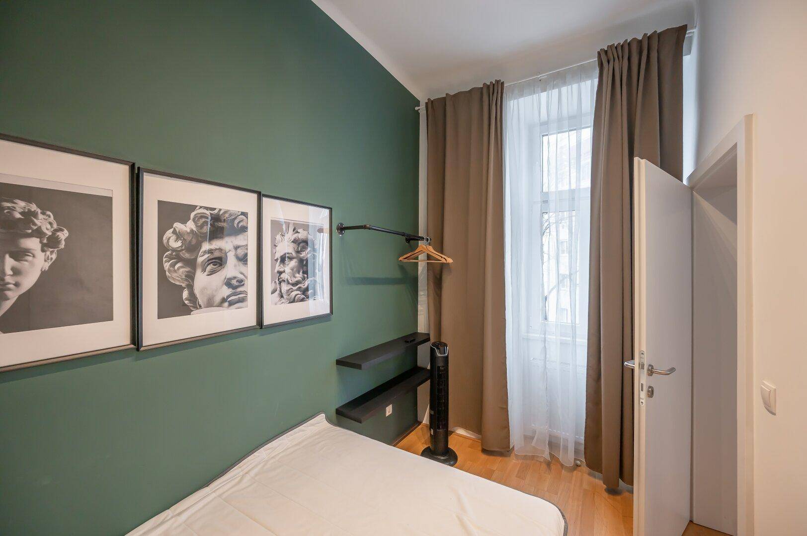 Gemütliches Schlafzimmer mit grüner Wand, Kunstwerken und Fenster mit Vorhängen.