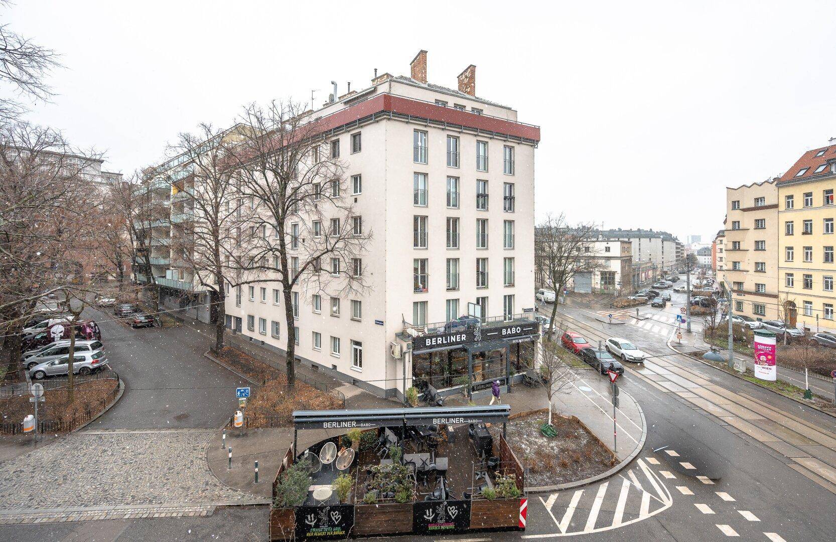 Eckgebäude in einer städtischen Umgebung mit Geschäften im Erdgeschoss und Straßenbahnlinien.