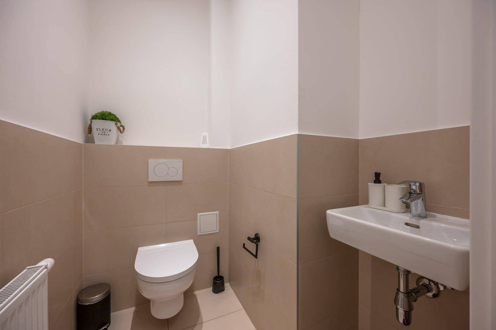 Kleines Gäste-WC mit Wand-WC, Waschbecken und beigefarbenen Fliesen an den Wänden.
