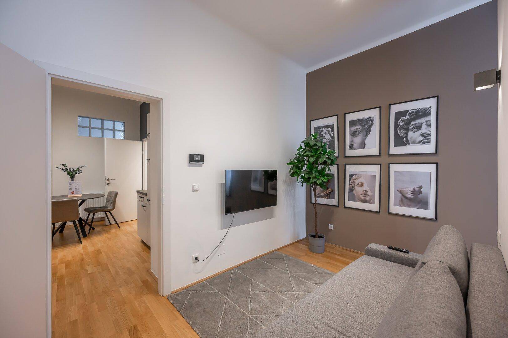 Wohnzimmer mit Sofa, Wandfernseher und Zugang zur Küche, hell und modern eingerichtet.