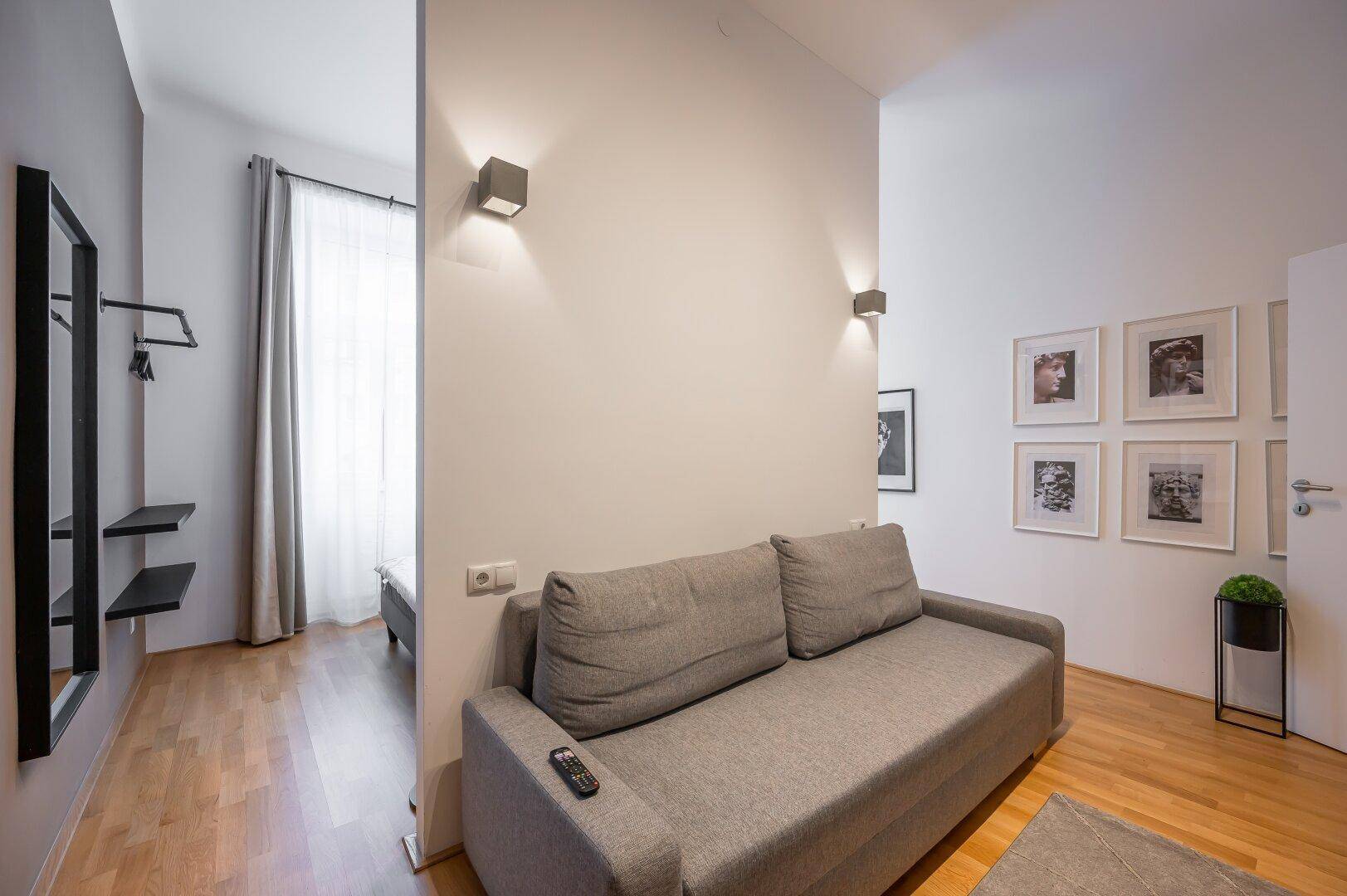 Heller Wohnbereich mit Sofa und Übergang zum Schlafzimmer, ausgestattet mit Holzboden.