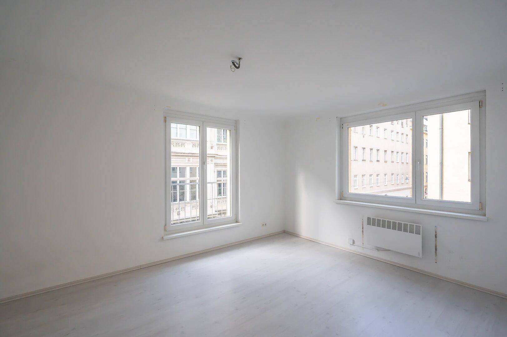 Geräumiges, helles Zimmer mit zwei großen Fenstern und weißem Laminatboden, ideal für Wohnzwecke.