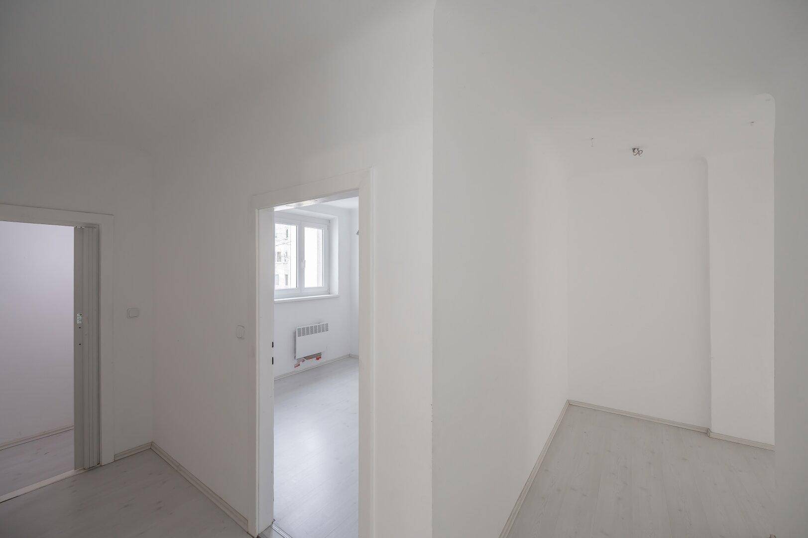 Heller Eingangsbereich mit Blick in ein weiteres Zimmer, gekennzeichnet durch weiße Wände und hellen Boden.