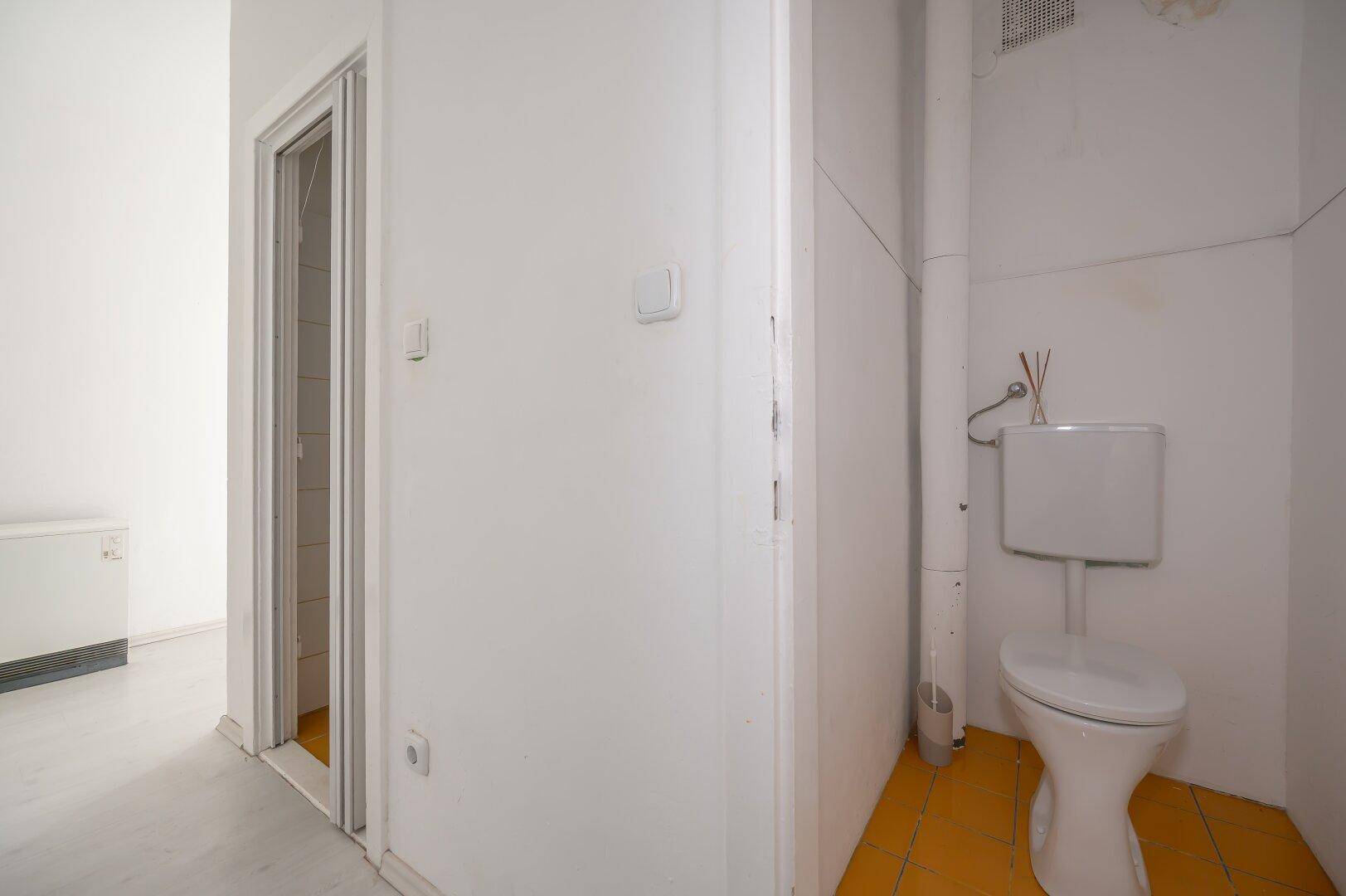 Separate Toilette mit gelben Bodenfliesen und einfacher Ausstattung, neben einem weiteren Raum.