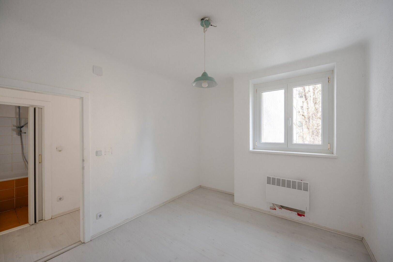 Kleineres, helles Zimmer mit Fenster und Pendelleuchte, angrenzend an ein Badezimmer.