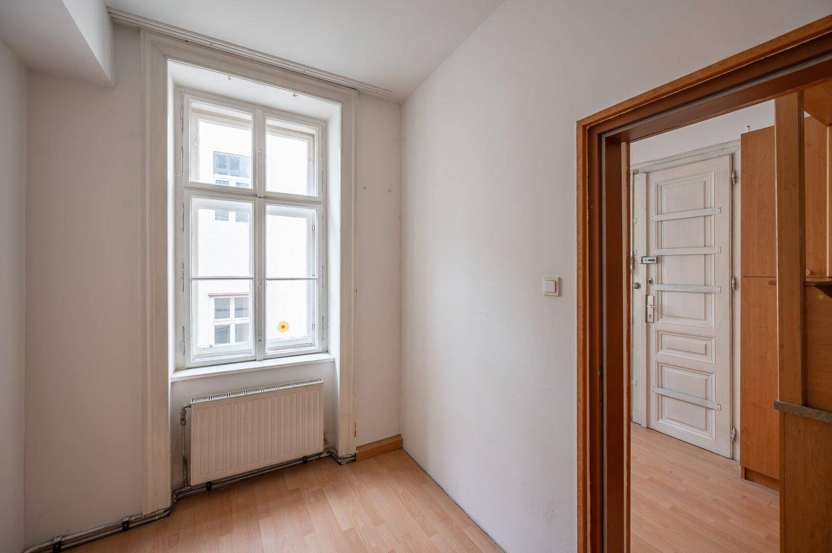 Kleiner Raum mit Fenster, Heizkörper und Holzboden, der zu einem weiteren Zimmer führt.